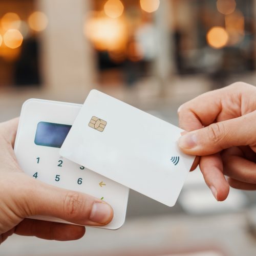 Unrecognizable customer with contactless white credit card paying to seller with terminal on blurred background of city street in daytime